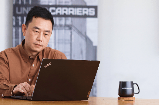 Man in brown shirt working on a ThinkPad laptop at a table with a black mug labeled xxx.